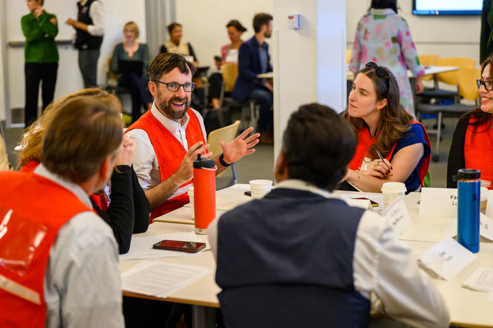 Event attendees mid-discussion, sitting at a table wearing bright vests